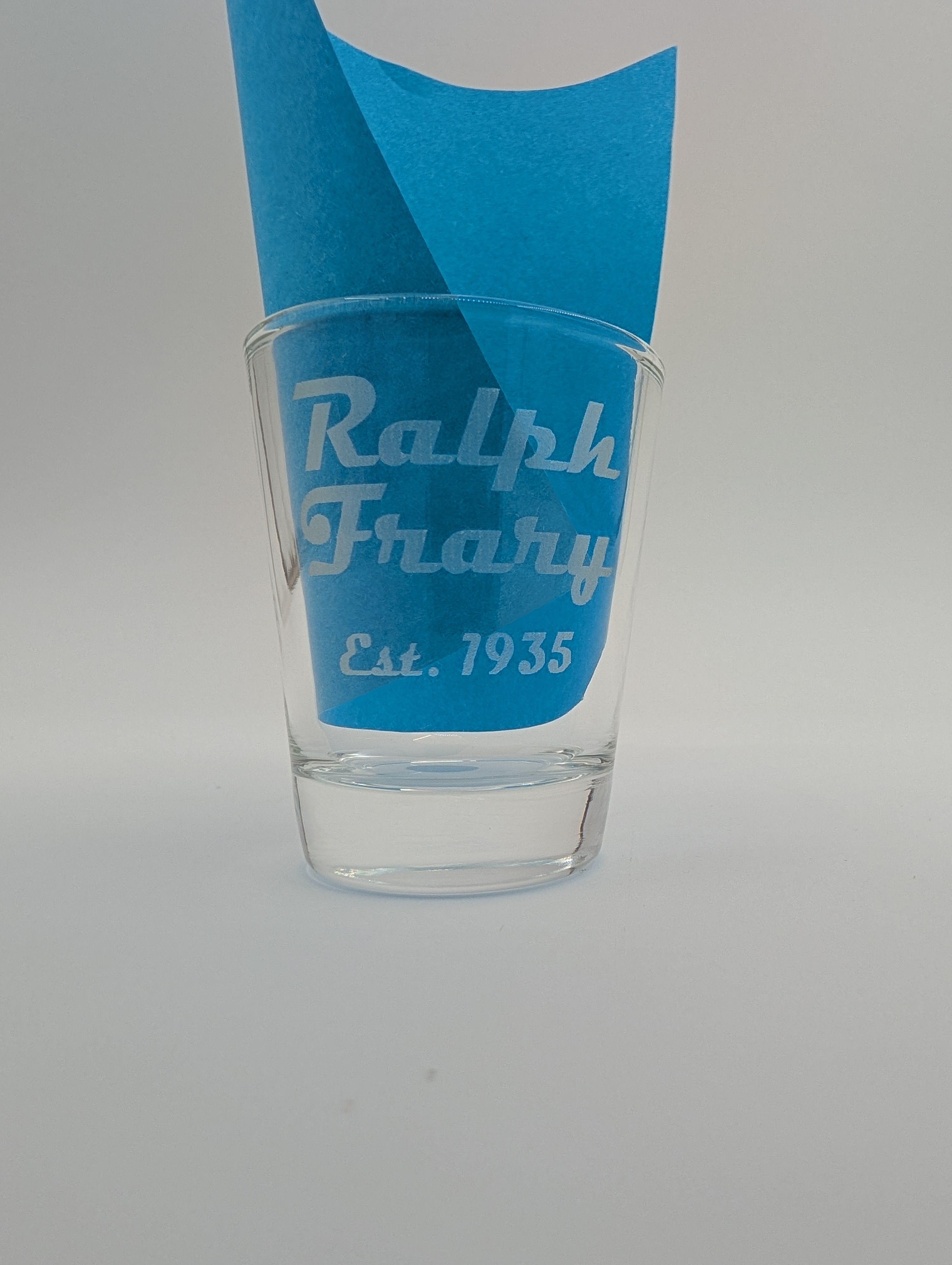 Clear glass shot glass with blue accent and 'Ralph Frary Est. 1935' text on a gray background